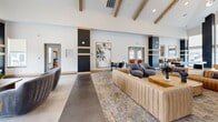 Located at 18905 E 59th Ave in Denver, CO, this single-floor community clubhouse offers a generously sized, thoughtfully designed gathering space that blends modern aesthetics with inviting comfort. The entrance leads into an expansive main living area distinguished by soaring vaulted ceilings with exposed wood beams, creating an airy, open atmosphere. Warm gold accents, dark charcoal column details, and rich wood trim run throughout, providing a cohesive and sophisticated design language. The primary lounge features plush sectional seating, curved accent chairs in soft grays and warm caramel tones, and a patterned area rug with gold undertones. A sleek fireplace with a wall-mounted TV anchors one end of the room, while large roller-shaded windows flood the space with natural light and offer views of the surrounding neighborhood. Statement pendant and chandelier lighting add an elegant finishing touch overhead. A secondary lounge area provides a more intimate setting, with rounded leather barrel chairs arranged around a compact coffee table, accompanied by casual high-top seating along the perimeter. The games area is outfitted with a full-size pool table and a shuffleboard table set on polished concrete floors, complemented by bistro-style seating and framed mirror accents. Together, these spaces form a well-rounded, amenity-rich environment ideal for social gatherings and everyday relaxation.