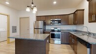This single-floor unit offers a thoughtfully designed open-concept layout that connects the kitchen and living area seamlessly. The kitchen is a standout feature, equipped with rich dark-stained cabinetry reaching toward the ceiling, speckled granite countertops, and a full suite of stainless steel appliances including a refrigerator, electric range, over-the-range microwave, and dishwasher. A granite-topped island adds additional prep and counter space, while a decorative chandelier overhead provides warm ambient lighting. Light wood-look flooring extends throughout the main living areas, complementing the neutral wall tones. The spacious living room is fitted with a ceiling fan and opens directly to a private balcony through a sliding glass door, offering outdoor views. The bedroom features plush carpet flooring and a large window with blinds that admits natural light and outdoor scenery. The bathroom is clean and functional, complete with a full soaking tub and shower combination, wood-look tile flooring, and a vanity with dark wood cabinetry. A dedicated laundry room provides added convenience, housing a full-size top-load washer and dryer alongside wire shelving for storage. The overall ambiance is fresh, modern, and move-in ready.