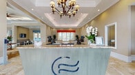 Located at 906 Oasis Palm Circle in Ocoee, Florida, this single-story clubhouse serves as the welcoming hub of the Oasis Palm community. The space makes an immediate impression at its reception area, where a granite-topped front desk — accented by a decorative logo panel — sits beneath a coffered ceiling with an ornate chandelier and recessed lighting, setting an upscale, hospitality-inspired tone throughout.

Beyond the reception, the main lounge area showcases rich hardwood flooring, soaring coffered and tray ceilings with exposed wood beams, and an abundance of natural light streaming through large windows dressed with valances. Comfortable, eclectic seating arrangements invite relaxation, while a dramatic stacked-stone fireplace with a wall-mounted television serves as a striking focal point.

A separate seating area features travertine tile flooring and an intimate circular arrangement of patterned accent chairs around a glass-top table, adjacent to a workstation with computer access. The kitchen is well-appointed with warm wood cabinetry, a herringbone tile backsplash, stainless steel appliances, and a dining area with views of the outdoors. A granite bar counter with bar seating offers a casual spot to gather. Throughout, the design blends warm tones, vibrant accent colors, and refined finishes to create an inviting and functional community environment.