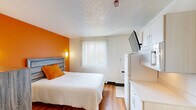 This Studio Deluxe in New Orleans, Louisiana, is a single-floor unit designed to maximize functionality within a compact footprint. The main living area serves as both a sleeping and living space, anchored by a bold orange accent wall that provides a vibrant focal point. A wood-panel headboard with matching dresser adds a rustic yet modern aesthetic, while wall-mounted sconces on either side of the bed offer warm, ambient lighting. Warm-toned laminate wood flooring runs throughout the unit, contributing to a cohesive and clean look.

The kitchenette area is efficiently arranged along one wall, featuring upper cabinets, a countertop workspace, a compact refrigerator, and a microwave. A wall-mounted flat-screen television is positioned above the refrigerator for convenient viewing. A window with vertical blinds allows natural light to fill the main space. Near the entry, a small dining table with chairs provides a dedicated space for meals, illuminated by a wall sconce.

The bathroom is cleanly appointed with wood-look flooring, a vessel sink set into a dark granite-style countertop, a large mirror, and chrome fixtures. Grab bars are installed throughout, adding accessibility. The textured ceiling is consistent across the unit, completing a straightforward, practical design.