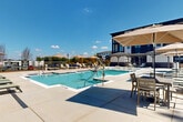 Located at 4980 Stonewall Tell Rd in Union City, Georgia, this outdoor amenity space is designed to deliver a resort-style experience for residents of the surrounding multi-story community. At the heart of the area is a generously sized swimming pool with crystal-clear water, a built-in tanning ledge, and stainless steel entry rails. The pool is framed by an expansive concrete deck outfitted with rows of cushioned lounge chairs, allowing ample space for relaxation under the Georgia sun. Large shade umbrellas and a covered pergola structure provide relief from the heat, while additional seating areas invite casual gathering and outdoor dining. The perimeter of the pool deck is enclosed by sleek wrought iron fencing, complemented by freshly landscaped mulch beds and young trees that add greenery and privacy to the setting. Towering pine trees line the far boundary, creating a natural backdrop that enhances the tranquil ambiance. The multi-story residential buildings surrounding the pool showcase a contemporary architectural design, combining brick, board-and-batten, and large-pane windows. The entire outdoor space benefits from abundant natural light and open skies, making it an inviting retreat that seamlessly blends modern design with natural surroundings.