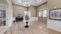 Located at 639 Preservation Trail in Webster, New York, this single-story community facility offers a thoughtfully designed interior that balances elegance with practicality. The entry lobby sets a refined tone with warm taupe walls, crisp white wainscoting, and wood-look flooring that extends throughout the space. A coffered tray ceiling with an ornate chandelier adds architectural distinction, while a central pedestal table with a succulent arrangement provides a welcoming focal point. Natural light filters through paneled windows framed in white trim, complementing the bright, airy atmosphere. A coffee station along one wall adds a convenient, hospitality-oriented touch near the main entrance.

A transitional corridor connects the lobby to additional areas, including a light-filled passage with a seating bench and multiple windows offering views to the exterior. The main gathering room is a standout space, featuring vaulted ceilings with a gabled roofline, a shiplap accent wall in slate gray, and a fireplace with a wood mantle as the room's centerpiece. A chandelier echoing the lobby's fixture anchors the dining area, where a large wooden table and black chairs are arranged atop a patterned area rug. Generous windows bring in natural light and exterior greenery.

Accessible restrooms are also provided, featuring tile flooring, dual vanities with dark countertops, and ADA-compliant fixtures, ensuring the facility meets the needs of all visitors.