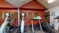 This fitness center is housed in a single-floor space defined by striking vaulted ceilings and bold terracotta-red walls that create an energizing atmosphere throughout. The room is illuminated by a combination of overhead fluorescent lighting and natural light streaming through multiple windows fitted with wood-framed blinds, including angled clerestory windows that follow the roofline and enhance the sense of height and openness. A ceiling fan provides additional air circulation across the space.

The cardio zone features Matrix-brand elliptical trainers and treadmills, positioned alongside large floor-to-ceiling mirrors that visually expand the area and allow users to monitor their form during workouts. A multi-station cable weight machine by TuffStuff serves as the centerpiece of the strength training area, accompanied by additional equipment including an inversion table and exercise balls.

A dedicated reading and relaxation corner adds a unique dimension to the fitness center, furnished with rolling bookshelves stocked with a wide selection of books. Wall-mounted flat-screen televisions are positioned for easy viewing from various points in the room, catering to both workout and leisure needs. Carpeted flooring covers much of the space, with rubberized mats placed beneath the heavier equipment. A water cooler is conveniently accessible, rounding out the practical amenities of this versatile, multipurpose fitness facility.