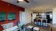 This single-floor residence showcases a thoughtfully designed open-concept layout that seamlessly connects the living, dining, and kitchen areas into one cohesive and functional space. The living area is anchored by a striking deep red accent wall, which adds warmth and personality to the room, complemented by a ceiling fan overhead and tasteful artwork. The space is furnished with a comfortable sofa adorned with patterned and solid accent pillows, alongside a coffee table arrangement that creates an inviting gathering area. Rich hardwood-style flooring flows throughout, grounding the entire space with a warm and cohesive aesthetic. The kitchen is a standout feature, boasting dark cabinetry, stainless steel appliances, and a breakfast bar with high-top seating — ideal for casual dining or entertaining. Elegant pendant lights hang above the bar counter, providing both functional and ambient illumination. Adjacent to the kitchen, a pass-through design enhances the open feel and promotes easy interaction between spaces. Multiple doorways lead to additional rooms, including what appears to be a laundry or utility closet, offering practical storage solutions. The overall ambiance is modern and inviting, with a bold yet balanced color palette that combines deep reds, neutral whites, and rich dark wood tones.