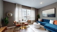 This single-floor residence offers a thoughtfully designed layout that maximizes both comfort and functionality. The entryway hallway sets the tone with warm wood-look flooring that carries throughout the space, neutral gray walls, and recessed lighting overhead.

The open-concept living and dining area is the heart of the unit, bathed in natural light through large windows dressed with sheer patterned curtains. The windows frame views of the surrounding urban streetscape, lending the space a connected, city-inspired feel. A compact dining nook with a round pedestal table and leather-accented chairs sits alongside a comfortable lounge area.

The kitchen is sleek and modern, featuring white upper cabinetry, gray lower cabinetry, white countertops with subway tile backsplash, and a full suite of stainless steel appliances including a gas range, microwave, dishwasher, and refrigerator. Recessed lighting illuminates the workspace efficiently.

The bedroom is a calm retreat, finished in soft neutral tones with wood flooring, ample wall space, and room for a full-size bed and bedside furnishings. The bathroom is bright and clean, featuring light tile flooring, a glass-enclosed shower, a vanity with storage, and crisp white walls that contribute to an airy, spa-like atmosphere.