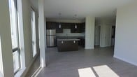 This single-floor space showcases a clean, contemporary design that emphasizes openness and functionality. The heart of the layout is a well-appointed kitchen featuring rich dark cabinetry, granite countertops, and a matching center island that provides additional workspace and casual seating potential. Stainless steel appliances, including a full-size refrigerator, microwave, and range, complement the modern aesthetic. Pendant lighting hangs above the island, adding focused illumination and a stylish touch to the space. The kitchen flows seamlessly into an expansive open living area, creating a connected, airy feel ideal for both everyday living and entertaining. Wide-plank gray hardwood-style flooring runs throughout, grounding the space with a neutral, contemporary tone. Large windows along the perimeter flood the interior with abundant natural light, casting warm sunlight across the floors and highlighting the clean lines of the space. Recessed lighting in the ceiling supplements the natural light for consistent brightness at all hours. Structural columns are thoughtfully integrated into the layout, adding architectural character without interrupting the open flow. A hallway visible toward the rear leads to additional rooms, offering a sense of depth and privacy beyond the main living area.