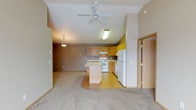 This single-floor home offers an open-concept design that connects the living area and kitchen in one cohesive space. The vaulted ceiling adds a sense of height and airiness, complemented by a ceiling fan that enhances comfort throughout. A pendant light fixture defines the dining or living zone, while natural-toned carpeting in the living area transitions smoothly to hardwood-style flooring in the kitchen, clearly delineating each functional space.

The kitchen is well-appointed with warm honey-toned wood cabinetry, a central island with a light countertop that provides additional workspace and casual seating potential, and a full suite of white appliances including a range, dishwasher, and refrigerator. An overhead range hood and under-cabinet lighting add practical utility. The layout is efficient, with ample counter space and storage cabinetry extending to the upper walls.

The neutral, taupe-toned walls throughout create a warm and inviting atmosphere. A doorway visible to the side suggests additional rooms beyond, offering private spaces separate from the main living areas. Overall, the space balances functionality and comfort in a clean, accessible single-level layout with a welcoming, understated aesthetic.
