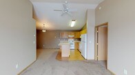 This single-floor home offers an open-concept design that connects the living area and kitchen in one cohesive space. The vaulted ceiling adds a sense of height and airiness, complemented by a ceiling fan that enhances comfort throughout. A pendant light fixture defines the dining or living zone, while natural-toned carpeting in the living area transitions smoothly to hardwood-style flooring in the kitchen, clearly delineating each functional space.

The kitchen is well-appointed with warm honey-toned wood cabinetry, a central island with a light countertop that provides additional workspace and casual seating potential, and a full suite of white appliances including a range, dishwasher, and refrigerator. An overhead range hood and under-cabinet lighting add practical utility. The layout is efficient, with ample counter space and storage cabinetry extending to the upper walls.

The neutral, taupe-toned walls throughout create a warm and inviting atmosphere. A doorway visible to the side suggests additional rooms beyond, offering private spaces separate from the main living areas. Overall, the space balances functionality and comfort in a clean, accessible single-level layout with a welcoming, understated aesthetic.