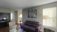 This single-floor residence offers a comfortable and cohesive living environment with a modern yet approachable aesthetic. The living area serves as the heart of the space, centered around a plush purple leather sofa that adds a bold pop of color against the cool gray accent wall. A coordinating patterned armchair and a sleek glass coffee table — adorned with fresh pink tulips — complete the seating arrangement, creating an inviting atmosphere for relaxation and conversation. A triptych of nature-inspired wall art featuring tree canopy imagery brings an organic, artistic touch to the main wall, enhancing the room's visual depth. Warm hardwood flooring runs throughout, grounding the space with richness and continuity. Natural light filters in through window treatments, while a floor lamp provides warm ambient lighting. The layout flows naturally toward additional areas of the home, including what appears to be an entryway with a classic paneled door, suggesting a well-connected and functional floor plan. The overall color palette — combining gray, cream, and warm wood tones — creates a calm and polished ambiance that is both livable and stylish.