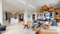 This thoughtfully designed single-floor space showcases a mid-century modern aesthetic carried consistently throughout each area. The open layout connects several distinct zones, all unified by wide-plank light oak flooring, crisp white walls, and carefully curated furnishings.

The main lounge area serves as the heart of the space, featuring a statement fireplace framed by geometric patterned tile and built-in shelving. Overhead, woven rattan pendant fixtures cast a warm glow over a seating arrangement of cognac leather chairs and a deep navy sofa. Large floor-to-ceiling sliding glass doors lead to an outdoor terrace, flooding the interior with natural light and offering urban rooftop views.

The kitchen is well-appointed with slate-grey cabinetry, marble-style tile backsplash, stainless and matte black appliances, and a generously sized terrazzo island topped with a globe pendant light cluster. Patterned accent wallpaper adds visual character to an adjacent area.

The dining space features bold geometric wallpaper as a focal backdrop, anchored by a round dark wood table, mid-century dining chairs, and amber glass pendant lighting. Throughout, the design balances refined detail with an approachable, welcoming ambiance.