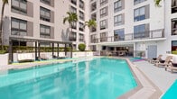 Located at 1642 NW 16th Street in Miami, FL, this amenity deck is a well-appointed outdoor retreat set within a modern residential high-rise. The centerpiece of the space is a generously sized resort-style swimming pool with crystal-clear turquoise water, framed by tall swaying palm trees and clean white architectural facades. Surrounding the pool, vibrant multi-colored striped flooring in teal, magenta, purple, and yellow-green adds a bold, energetic aesthetic that reflects Miami's dynamic design culture. Sun loungers are positioned along the pool deck, offering residents comfortable spots to relax and enjoy the Florida sunshine. A covered lounge area provides a shaded retreat, outfitted with contemporary wire-frame furniture, a wall-mounted flat-screen TV, and open-air views overlooking the pool and the Miami skyline beyond. Adjacent to the lounge, an interior amenity room features a dining table with seating and access to a kitchenette, ideal for social gatherings. A perimeter walkway enclosed by metal mesh railings offers sweeping panoramic views of the surrounding Miami cityscape under open skies. Lush greenery and manicured planters throughout add a refreshing natural contrast to the modern concrete and steel architecture, creating a balanced, resort-like atmosphere.