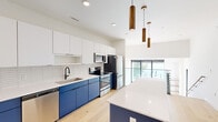 This contemporary single-floor residence in Oshkosh, Wisconsin offers a thoughtfully designed layout with one bedroom, one full bath, and a half bath. The open-concept kitchen is a standout feature, showcasing bold navy blue lower cabinetry paired with crisp white upper cabinets and a matching white quartz countertop. A textured white elongated tile backsplash adds dimension, while a large center island provides additional workspace and seating. Stainless steel appliances, including a range, microwave, and refrigerator, complete the modern aesthetic. Warm wood pendant lights hang above the island, adding an inviting contrast to the clean palette. Light hardwood flooring extends throughout the living areas, contributing to a bright and airy atmosphere. The entryway makes a memorable first impression with a custom slatted wood and navy partition wall paired with built-in cabinetry, framing the space with architectural interest. The bedroom is bathed in natural light from expansive black-framed windows that frame serene waterfront views. The half bath features a navy vanity with matte black fixtures and a large wall mirror. A dedicated laundry closet with a stacked washer and dryer adds everyday convenience to this well-appointed residence.