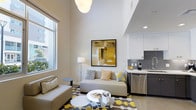 This thoughtfully designed two-floor space makes a striking impression with its soaring ceilings that amplify the sense of openness and volume throughout the main living area. Large windows flood the space with natural light, enhancing the clean, neutral palette of white walls and light finishes that create a fresh, airy atmosphere.

The open-plan living area is anchored by a comfortable sofa set arranged around a sleek round coffee table, accented by a bold geometric patterned rug in yellow, grey, and white that adds a vibrant focal point to the room. A large abstract artwork in warm golden tones adorns the wall, complementing the color scheme and adding an artistic touch to the space.

The kitchen flows seamlessly from the living area and features contemporary white cabinetry, a distinctive grey mosaic tile backsplash, and stainless steel appliances, offering both functionality and style. Under-cabinet lighting and clean countertops contribute to the kitchen's polished, modern look.

A pendant light and floor lamp provide layered lighting options, allowing the ambiance to be easily adjusted. The two-floor layout adds a dynamic vertical dimension to the space, making it feel expansive while maintaining an intimate and welcoming character throughout.