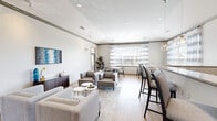 Located at 1200 Belfast Drive in Saint Peters, Missouri, this well-appointed community amenity space is thoughtfully designed across a single floor. The open-concept lounge area serves as the centerpiece, featuring warm light hardwood flooring, recessed lighting, and large windows dressed with sheer striped curtains that invite abundant natural light. Comfortable gray sofas and armchairs are arranged around low coffee tables, creating an inviting setting for socializing or relaxing. A granite-topped bar counter with pendant globe lighting separates the lounge from a kitchen area equipped with dark wood cabinetry, a tiled backsplash, wall-mounted TV, and full-size black refrigerator. A casual dining nook adjacent to expansive windows provides a bright spot for informal gatherings, furnished with a round wood table and modern wishbone chairs. A dedicated business center offers dual computer workstations with a shared printer, accented by coffee-themed artwork. The fitness room is outfitted with a cable weight machine, elliptical, and workout bench beneath a ceiling fan and recessed lighting. A leasing or information area with a literature display wall and curated artwork rounds out the space, reflecting a cohesive, contemporary aesthetic throughout.