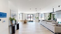 Located at 397 Millburn Ave in Millburn, NJ, this thoughtfully designed single-floor space offers a generous footprint across two living areas, a dining room, kitchen, and bedroom. The open-concept main living and dining area is bathed in natural light through floor-to-ceiling black-framed windows dressed with sheer curtains, creating an airy, welcoming atmosphere. Light-toned hardwood floors extend throughout, complementing the clean white ceilings with recessed lighting. A second living area features a striking marble-surround linear fireplace with a mounted display above, plush patterned carpeting, and bold decorative mirrors with sculptural frames, lending a contemporary, curated aesthetic. The dining room is a standout, showcasing a textured accent wall in deep blue-grey tones, a long communal table beneath a striking gold-and-black linear chandelier, and a sliding barn door detail. A dedicated game room nook with large windows overlooking the outdoor terrace adds a recreational dimension. Outside, a spacious tiled terrace provides seating areas with umbrellas and lounge furnishings, surrounded by views of the modern brick-and-panel building exterior with manicured landscaping below. The overall ambiance is polished, modern, and community-oriented.