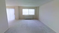 This single-floor space offers a clean, functional layout centered around a spacious, open living area. The room is finished with plush neutral-toned carpeting that extends throughout, providing a soft and comfortable underfoot feel. A large window dressed with vertical blinds serves as the focal point of the main living space, allowing an abundance of natural light to fill the room and creating a bright, airy atmosphere. A baseboard heating unit is positioned beneath the window, ensuring efficient climate control throughout the colder months. The walls are finished in a crisp, neutral tone that complements the light-colored carpet, lending the space a clean and versatile aesthetic well-suited for a variety of furnishing styles. A secondary access point with vertical blinds is visible to the side, suggesting an additional source of natural light or outdoor access. The overall ambiance is simple and understated, offering a blank canvas that can be easily personalized. The layout feels open and unobstructed, making the space feel roomy and accommodating despite its straightforward design.