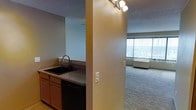 This single-floor unit features an open-concept layout that connects the kitchen and living area, creating a cohesive and functional living space. The kitchen is positioned along one wall and includes warm wood-toned cabinetry, a black undermount sink with a modern faucet, a dark countertop, and a black dishwasher. A structural column separates the kitchen from the adjoining living area, adding architectural definition while maintaining visual openness. Track lighting mounted overhead provides direct illumination to the kitchen workspace. The textured ceiling adds a classic finishing detail throughout the space. The living area features wall-to-wall carpeting, lending a comfortable and soft underfoot feel. A large floor-to-ceiling window with vertical blinds allows abundant natural light to fill the room and provides an expansive outdoor view. A baseboard heating unit is positioned beneath the window, offering efficient climate control. The neutral color palette of beige and cream tones throughout creates a calm, inviting atmosphere well-suited for a variety of interior design styles. The overall layout emphasizes practicality and light-filled living.