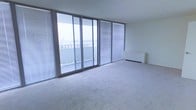 This single-floor space is defined by its striking wall of floor-to-ceiling sliding glass doors, which span the width of the main living area and flood the interior with abundant natural light. The doors open directly onto a private balcony, offering expansive views of the surrounding cityscape — a standout feature that brings an elevated, open-air quality to the space. Horizontal venetian blinds are fitted throughout, providing flexible light and privacy control throughout the day. The living area is finished with soft wall-to-wall carpeting in a neutral tone, contributing to a calm and comfortable ambiance. The walls and ceiling are rendered in a clean, off-white palette, enhancing the sense of brightness and spaciousness. A baseboard heating unit is positioned along one wall, ensuring year-round comfort. The overall design is minimal and functional, with an open layout that maximizes the connection between the interior and the outdoor balcony space. The combination of generous glazing, city views, and a neutral interior finish makes this a well-lit, versatile space suited to a variety of uses.