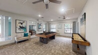 This single-floor recreational space is designed as a contemporary game and social lounge, offering a well-appointed environment for leisure and entertainment. The main area is anchored by a full-size pool table with a sleek black felt surface and a warm wood-and-dark-metal frame, complemented by a shuffleboard table along the adjacent wall. Patterned carpet tiles in neutral tones cover the main floor, lending texture and a modern aesthetic throughout.

Multiple ceiling fans are distributed across the space, ensuring comfortable airflow, while recessed lighting provides consistent, even illumination. Large windows along multiple walls invite generous natural light and offer views of the surrounding greenery, enhancing the open feel of the room.

A lounge nook features a wall-mounted television, a tufted accent chair with a teal pillow, and a small side table, set against a bold teal accent wall that adds a vibrant pop of color. Framed abstract artwork in teal and neutral tones is displayed throughout, reinforcing the cohesive modern design scheme. A wall-mounted cue rack keeps equipment organized and accessible.

The space connects to an adjacent room visible through a large mirror or pass-through, extending the sense of openness. Multiple exterior doors provide convenient access to the outdoors, making this a functional and inviting communal gathering area.