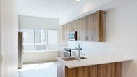 This well-designed multi-floor home offers four bedrooms, two bathrooms, and a thoughtfully arranged layout spread across three levels. The ground floor includes an attached garage with ample space and utility access, alongside additional storage areas. A carpeted staircase with a sleek metal railing connects the levels, providing a clean transition between floors. The main living level features a combined kitchen and living area bathed in natural light from large windows. The kitchen is appointed with warm wood-toned cabinetry extending to the ceiling, white quartz countertops, stainless steel appliances including a built-in microwave, electric range, dishwasher, and refrigerator, and a peninsula with a double undermount sink — a functional and stylish centerpiece. Light hardwood flooring flows throughout the main living spaces, enhancing the bright, contemporary ambiance. The bathrooms feature crisp white subway-tiled tub-shower combinations with modern matte black fixtures and accented tile flooring. The generously sized primary bedroom, the largest of the four, opens directly onto a private balcony through sliding glass doors, offering outdoor views of surrounding greenery. Recessed lighting throughout maintains a clean, modern aesthetic across the entire home.