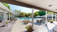 Located at 3750 Carothers Pkwy in Franklin, Tennessee, this single-story amenity facility serves as a resort-style outdoor retreat. The building showcases classic Southern architecture with gray horizontal lap siding, crisp white trim, white columns, and a welcoming covered front porch. Double glass-panel doors and large transom windows allow ample natural light into the interior while providing views of the surrounding grounds.

A wide wraparound deck extends along the exterior, finished with rich hardwood-toned decking and lined with black metal railings. The deck is furnished with comfortable seating and decorative potted flowers, creating an inviting transitional space between indoors and outdoors. Ceiling-mounted light fixtures illuminate the covered porch, which runs the full length of the building.

The centerpiece of the property is a large, rectangular resort-style swimming pool with crystal-clear blue water. The expansive concrete pool deck is outfitted with rows of lounge chairs and blue-accented seating, ideal for relaxing in the sun. Patio umbrellas provide shaded areas throughout the deck. A separate covered gazebo structure with additional seating is positioned poolside, offering a shaded gathering spot.

Mature trees and lush greenery frame the entire property, providing privacy and a serene natural backdrop. The surrounding grounds include open patio space accommodating outdoor dining and social areas.