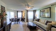 This single-floor space offers a thoughtfully designed open-concept layout that integrates the living and dining areas into one cohesive, welcoming environment. Large windows dressed with elegant dark curtains and white blinds flood the space with natural light, creating an airy and inviting atmosphere throughout.

The living area features a soothing sage green accent wall that adds a touch of color and warmth. A ceiling fan with a built-in light fixture serves as a functional centerpiece overhead, complementing the textured ceiling. The space is well-appointed with a neutral-toned sofa, a tufted leather ottoman as a coffee table, and a patterned area rug that anchors the seating arrangement.

The dining area, seamlessly connected to the living space, accommodates a dark wood dining table with matching upholstered chairs, offering a comfortable space for meals and gatherings. Track lighting along the ceiling near the dining area provides additional illumination.

The overall ambiance is warm and comfortable, with a neutral color palette accented by soft greens and rich dark tones. The open flow between areas, combined with generous window placement, enhances the sense of spaciousness and makes this a bright, functional, and welcoming home.