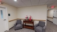 This single-floor facility is thoughtfully designed to provide a comfortable and functional environment for its occupants. The lounge area sets a warm, welcoming tone with plush neutral-toned sofas, a patterned accent chair, and a rustic wooden coffee table arranged around a patterned carpet. Wall art and decorative greenery enhance the relaxed atmosphere, while windows allow natural light to filter in beneath the suspended tile ceiling.

Adjacent to the lounge, the dining area accommodates multiple wooden dining tables and chairs, offering ample seating for group gatherings. Framed artwork adorns the walls, and the space transitions seamlessly into a kitchenette outfitted with light wood cabinetry, a countertop with a sink, a full-size refrigerator, and small appliances including a coffee maker. The kitchen extends into a well-lit hallway with tile flooring that connects the various areas of the facility.

The restrooms are finished to a high standard, featuring earthy stone-look tile covering both the floors and lower walls, grab bars for accessibility, and tasteful decorative accents. The overall design of the space balances practicality with a homey aesthetic, making it well-suited for community use, group activities, or supportive care settings.