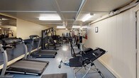 This fitness center offers a comprehensive workout environment spread across a single floor. The space is outfitted with durable, easy-to-clean flooring throughout and is illuminated by bright fluorescent lighting, ensuring a well-lit atmosphere for exercise at any time of day. Overhead ceiling fans help circulate air and maintain a comfortable environment during workouts.

The cardio section is equipped with multiple treadmills, a recumbent bike, and an elliptical trainer, providing ample options for aerobic exercise. The strength training area features a Weider Pro 4900 multi-station home gym, a dedicated bench press station with free weights and a barbell rack, and an inversion table for core and back work. Additional cable and resistance machines line the walls, offering a variety of targeted muscle exercises.

A large wall mirror in the strength training area allows users to monitor their form. The space is accessed via a carpeted staircase with a clearly marked exit sign, and a secondary door provides an additional entry and exit point. The textured ceiling and paneled wall sections give the space a functional, no-frills character well-suited to its purpose as a practical, fully equipped fitness facility.