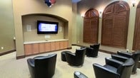 This single-floor media lounge offers a well-appointed gathering space designed for comfort and entertainment. At the heart of the room, a large wall-mounted flat-screen television is set within a recessed arched niche, flanked by warm wood built-in cabinetry that provides ample storage below. The seating arrangement features multiple black leather swivel chairs positioned to face the screen, creating an inviting and functional viewing environment. The room's warm tan and beige walls contribute to a relaxed, welcoming ambiance, while decorative wall sconces provide soft accent lighting throughout. A standout architectural feature is the pair of tall arched windows dressed with rich, dark-stained wooden louvered shutters, adding a classic and elegant touch to the space. The neutral-toned carpeted flooring ties the room together, enhancing its cozy atmosphere. The combination of thoughtful design elements — from the arched architectural details to the custom millwork and leather furnishings — makes this space an ideal setting for group viewings, casual gatherings, or simply unwinding in a stylish and well-designed environment.
