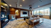 Located at 8320 University Executive Park Drive in Charlotte, NC, this single-floor lounge space offers a thoughtfully designed interior that blends contemporary aesthetics with warm, natural materials. The open layout is organized into several distinct functional zones, each contributing to a cohesive yet varied atmosphere throughout.

The main lounge area features rich wood-paneled accent walls contrasted against textured white wall surfaces, creating a sophisticated visual balance. A wall-mounted flat-screen television serves as a focal point, flanked by curated wall art, including a striking natural wood slab piece. Plush, neutral-toned sectional seating is anchored by a live-edge wood coffee table, while a dramatic cylindrical pendant light fixture adds an architectural element overhead. Large windows along one wall admit generous natural light and offer outdoor views.

A casual dining area includes a live-edge communal table with mixed woven and upholstered seating beneath an artistic cluster pendant light. Bold abstract artwork in teal, yellow, and white is displayed against deep navy accent walls, adding strong visual interest. Smaller café-style tables near the windows provide additional flexible seating.

A more intimate nook features a deep teal geometric tile accent wall, open dark shelving with curated décor, and a high-back lounge chair, offering a quieter retreat within the space. Throughout, patterned area rugs unify the zones cohesively.