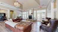 This thoughtfully designed single-floor space spans a generous footprint, encompassing multiple living areas, a combined dining and living room, and a dedicated office. The main living area commands immediate attention with its soaring vaulted ceiling, an eye-catching globe chandelier, and a central fireplace framed by tall, multi-pane windows and French doors that flood the room with natural light and offer views to the outdoors. Rich hardwood-style flooring flows throughout, anchoring the warm, neutral color palette. Classic tufted leather sofas and wingback chairs lend a sophisticated yet welcoming ambiance. A secondary living area provides a more relaxed retreat, featuring a built-in white cabinetry unit with a mounted television, large windows with greenery views, and layered area rugs for added comfort. The combined dining and living room offers an airy, light-filled setting with oversized windows and a round dining table suited for intimate gatherings. The private office is equipped with built-in wood cabinetry, a workstation, and a tall window overlooking mature trees, creating a bright and functional workspace. Throughout, the space maintains a cohesive, transitional aesthetic with carefully chosen furnishings, abundant natural light, and a calm, inviting atmosphere.