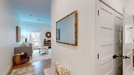 Located at 27 Park Rd in West Hartford, Connecticut, this single-floor residence offers a thoughtfully designed open-concept layout with a clean, contemporary aesthetic. Light-toned hardwood flooring flows throughout the space, complementing the crisp white walls and creating a warm, cohesive atmosphere.

The entry area features a panel-style door and a framed mirror, providing a welcoming transition into the main living areas. The open living and sleeping space is generously proportioned, with large windows on multiple walls that invite abundant natural light and offer views of the surrounding greenery and neighboring architecture. The versatile layout accommodates both a comfortable lounge area and a sleeping area within the same open footprint.

The kitchen is efficiently designed with floor-to-ceiling gray cabinetry, white countertops, and a slate-blue subway tile backsplash. Stainless steel appliances — including a range, microwave, dishwasher, and refrigerator — provide a polished, modern look. A small bar-height counter offers additional seating.

A hallway connects to additional areas, including what appears to be a bathroom and closet space. Throughout the unit, the neutral palette, recessed lighting, and roller shades contribute to a bright, modern, and functional living environment.