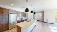 Located at 746 Element Way in Ashwaubenon, Wisconsin, this well-appointed single-floor residence combines contemporary design with practical finishes throughout. The heart of the home is an open-concept kitchen that immediately impresses with its large center island topped with a light quartz countertop and an undermount sink. Rich warm-toned cabinetry lines the walls, complemented by a full suite of stainless steel appliances including a refrigerator, electric range, and microwave. Three sleek black pendant lights hang above the island, adding a modern touch, while light-toned hardwood-style flooring extends through the main living areas. Natural light flows in through generously sized windows, offering pleasant views of surrounding autumn foliage. The bedrooms feature soft carpeting and large windows that bring in abundant daylight; one bedroom includes a ceiling fan for added comfort. Two bathrooms serve the residence — one with a walk-in shower and marble-style tile flooring, and another with a full soaking tub/shower combination and coordinating finishes. A dedicated laundry room houses a Whirlpool front-loading washer and dryer with built-in wire shelving above. The overall aesthetic is clean, neutral, and move-in ready.