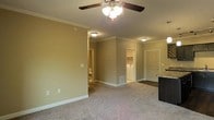 This single-floor space features an open-concept layout that connects the main living area to the kitchen and utility spaces. The living area is finished with plush wall-to-wall carpeting and neutral taupe-toned walls, creating a warm and understated atmosphere. Crown molding runs along the ceiling perimeter, adding a refined architectural detail throughout. A ceiling fan with an integrated light fixture provides both ventilation and ambient lighting to the main living space, while flush-mount ceiling lights illuminate the connecting hallways and additional areas.

The layout reveals a dedicated laundry area with a washer and dryer visible through an open doorway, offering added in-unit convenience. An adjacent doorway leads to a bathroom, partially visible from the main living space. To the right, a dark-finished countertop marks the transition into the kitchen area, which features contrasting cabinetry. White-painted doors and trim throughout provide clean contrast against the neutral walls. Vinyl flooring is visible in the entryway and utility areas, transitioning to carpet in the main living space. The space also includes a floor vent for climate control and multiple electrical outlets positioned along the baseboards.