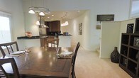 This single-floor residence showcases a thoughtfully designed open-concept layout that seamlessly connects the kitchen, dining, and living areas into one cohesive and functional space. The kitchen is a standout feature, equipped with rich dark cabinetry, dark granite countertops, stainless steel appliances, and a dedicated range hood, all illuminated by recessed lighting and a modern track light fixture overhead. A breakfast bar provides casual seating and further reinforces the open flow between the kitchen and adjoining spaces. The dining area is anchored by a substantial dark wood dining table complemented by matching chairs and a stylish multi-pendant light fixture that adds warmth and definition to the space. Neutral-toned carpeting and light walls create a clean, welcoming ambiance throughout, while decorative wall art adds subtle personality. Curved architectural wall details lend a soft, refined character to the interior. A staircase visible in the background hints at additional vertical space, while the overall layout is designed to encourage effortless movement and comfortable everyday living. The combination of layered lighting, quality finishes, and an open floor plan makes this a warm and inviting home.