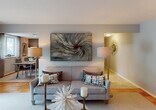 This thoughtfully designed single-floor residence offers a seamless blend of comfort and contemporary style. The open living room serves as the heart of the home, featuring neutral gray walls, warm wood-toned flooring, and plush carpeting underfoot. Bold artwork anchors the main wall, complementing the relaxed, curated aesthetic throughout.

Adjacent to the living area, the dining space features a striking accent wall in deep slate blue, providing a sophisticated backdrop for the dining table and sideboard. An open pass-through connects this area directly to the kitchen, which is outfitted with rich cherry wood cabinetry, granite countertops, a stainless steel sink, and a full suite of stainless steel appliances including a range, over-the-range microwave, and dishwasher.

The primary bedroom offers a tranquil retreat with a decorative accent wall, a substantial wood-panel headboard, and a window dressed with elegant draping curtains that allow natural light to filter in. The second bedroom is equally well-proportioned, featuring hardwood parquet flooring, a dark wood headboard, and bright windows with garden views.

The bathroom is clean and functional, featuring light tile flooring, a rich wood vanity with a white countertop, a framed mirror, and a tub-shower combination. Throughout the residence, a cohesive neutral palette and natural light create a calm, welcoming ambiance.