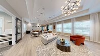 This single-floor clubhouse is thoughtfully designed to serve as both a social gathering space and a wellness facility. The living room is a refined lounge area that exudes modern elegance, anchored by a tray ceiling adorned with a striking multi-globe chandelier that casts warm, inviting light throughout the room. Large windows dressed with sheer white curtains and blinds line the walls, flooding the space with abundant natural light and offering views of the surrounding greenery. Light wood-look flooring runs throughout, complementing the neutral taupe wall tones. A classic white fireplace with a dark built-in shelving unit serves as a focal point, while a curved frosted-glass room divider with LED accent lighting adds a contemporary architectural detail that elegantly separates the lounge from the fitness area. The exercise room is a well-lit, practical space outfitted with professional-grade equipment, including a Star Trac treadmill, a cable weight machine, and a free weight rack. Natural light pours in through multiple windows, and a wall-mounted mirror enhances the sense of openness. Durable stone-style flooring provides a sturdy foundation for workouts. Together, these two spaces create a cohesive and versatile clubhouse environment.
