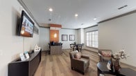 This single-floor community space is thoughtfully designed to serve multiple functions, blending a welcoming lounge, a dining area, and a fully equipped business center into one cohesive environment. Wide-plank hardwood-style flooring runs throughout, providing a warm and consistent foundation that ties each zone together seamlessly.

The lounge area features a comfortable arrangement of leather armchairs accented with patterned throw pillows, centered around a dark curved coffee table. Natural light filters in generously through plantation shuttered windows, while dark-trimmed entry doors with glass panels add an elegant architectural touch. Crown molding and recessed lighting contribute a refined, polished look to the neutral gray walls.

A dedicated dining nook offers a dark wood high-top table surrounded by sleek metal-frame chairs, set beneath framed contemporary artwork. Plantation shutters on the adjacent window allow for adjustable light control in this area.

The business center is anchored by a modern workstation counter finished in warm wood tones, accommodating multiple desktop computers with ergonomic task chairs. An eye-catching orange accent wall with decorative pendant lighting distinguishes this zone and adds a vibrant pop of color to the overall neutral palette. A wall-mounted flat-screen television completes the space, making it equally suited for both productivity and relaxation.