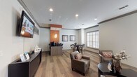 This single-floor community space is thoughtfully designed to serve multiple functions, blending a welcoming lounge, a dining area, and a fully equipped business center into one cohesive environment. Wide-plank hardwood-style flooring runs throughout, providing a warm and consistent foundation that ties each zone together seamlessly.

The lounge area features a comfortable arrangement of leather armchairs accented with patterned throw pillows, centered around a dark curved coffee table. Natural light filters in generously through plantation shuttered windows, while dark-trimmed entry doors with glass panels add an elegant architectural touch. Crown molding and recessed lighting contribute a refined, polished look to the neutral gray walls.

A dedicated dining nook offers a dark wood high-top table surrounded by sleek metal-frame chairs, set beneath framed contemporary artwork. Plantation shutters on the adjacent window allow for adjustable light control in this area.

The business center is anchored by a modern workstation counter finished in warm wood tones, accommodating multiple desktop computers with ergonomic task chairs. An eye-catching orange accent wall with decorative pendant lighting distinguishes this zone and adds a vibrant pop of color to the overall neutral palette. A wall-mounted flat-screen television completes the space, making it equally suited for both productivity and relaxation.