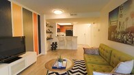 This single-floor space showcases an open-concept design that seamlessly connects the living area and kitchen, creating a cohesive and functional layout. The living area is anchored by light hardwood-style flooring, a bold black-and-white chevron area rug, and a tufted olive-green sofa that adds a striking pop of color. A round wooden coffee table serves as a central focal point, styled with decorative accents that enhance the eclectic aesthetic.

An accent wall featuring orange and dark vertical stripes introduces a dynamic visual element near the entryway, complemented by open wooden shelving displaying curated decorative pieces. The kitchen is positioned toward the rear of the space and features warm cherry-toned cabinetry, a black refrigerator, and light countertops. A peninsula with a butcher-block-style counter and two wire bar stools creates a casual dining and socializing area that bridges the kitchen and living space.

Ceiling-mounted lighting provides consistent illumination throughout, while the neutral wall tones balance the bolder design choices. The overall ambiance is modern, lively, and thoughtfully styled with a mix of warm tones and contemporary furnishings.