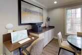 This single-floor business center is thoughtfully designed to support both focused work and collaborative meetings. The space features warm greige walls, crown molding, and durable wood-look flooring throughout, creating a polished yet welcoming professional atmosphere. Recessed lighting provides consistent, even illumination across the entire area.

A central meeting zone is anchored by a round glass-topped table surrounded by upholstered cream chairs, ideal for small group discussions or informal gatherings. Adjacent to this area, a built-in workstation features a rich dark wood desktop paired with crisp white cabinetry below, offering ample storage and surface space. The station is equipped with a desktop computer and multifunction printer, making it a fully functional work hub. A large framed abstract artwork is mounted above the cabinetry, adding a refined decorative touch.

A prominent wall-mounted whiteboard serves as a key collaborative tool, visible from multiple vantage points within the space. Natural light filters in through several windows fitted with horizontal blinds, offering views of the surrounding greenery. A glass-paneled exterior door further enhances the connection to the outdoors. A connecting corridor leads to additional areas beyond, suggesting this business center is part of a larger facility.