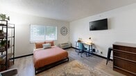 This single-floor studio is thoughtfully designed to maximize functionality within a compact footprint. The main living area seamlessly integrates sleeping, dining, and relaxing zones, anchored by light wood-style flooring that runs throughout the space. A full-sized bed serves as the centerpiece, accompanied by a dedicated workspace featuring a desk and seating, a wall-mounted flat-screen TV, and a decorative wall clock. Natural light filters in through a window fitted with horizontal blinds, brightening the room with an airy feel.

The kitchenette is equipped with dark cabinetry, a stainless steel full-sized refrigerator, a microwave, and a sink, offering essential cooking and food storage capabilities. A rolling utility cart provides additional counter and storage space, while open shelving above the cabinetry adds further organization. A compact dining area with a wooden high-top table and bar stool seating is conveniently positioned near the entrance corridor.

The bathroom is clean and well-organized, featuring a vanity with a granite-style countertop, a large full-length mirror, a towel bar, and a separate water closet with a toilet and additional towel storage. The textured ceilings and neutral white walls throughout contribute to a bright, functional, and cohesive aesthetic.