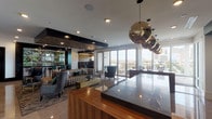 Located on Smith Street in Houston's vibrant 77006 corridor, this single-floor amenity space presents a refined, contemporary design that balances functionality with upscale aesthetics. Floor-to-ceiling glass walls and expansive windows flood the interior with natural light while framing sweeping views of the Houston skyline, creating a seamless connection between the indoor environment and the cityscape beyond.

The space is organized into several distinct zones. A central lounge area features polished large-format tile flooring, dark architectural columns, tufted seating, and accent chairs arranged around a reflective gold coffee table. Adjacent to this, a well-appointed kitchen area showcases rich walnut millwork, a substantial island with dark stone countertops, bar seating, and a dramatic iridescent tile backsplash, all illuminated by a striking cluster of spherical metallic pendant lights.

A dedicated conference or dining area positioned along the glass perimeter offers direct city views, complemented by the same bold pendant fixtures. A separate lounge nook features a slatted wood-paneled ceiling, a large sectional sofa, live-edge wood coffee tables, and a wall-mounted flat-screen display. A billiards table is also visible within the broader floor plan, rounding out the entertainment offerings. Throughout, the design palette of dark metals, warm woods, and stone creates a cohesive, sophisticated ambiance.