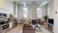 This thoughtfully designed single-floor unit is a striking example of historic adaptive reuse, where classical architectural grandeur meets modern living. The expansive open-plan living and dining area is the centerpiece of the space, defined by soaring ceilings, ornately detailed columns with decorative capitals, and tall arched windows that flood the interior with abundant natural light. A fireplace with a dark marble surround anchors the living area, complemented by a TV console, plush seating, and light hardwood flooring that extends throughout the unit.

The kitchen is efficiently laid out with white shaker cabinetry, granite countertops, a stainless steel appliance suite including a refrigerator, range, microwave, and dishwasher, and generous counter space. A dining area sits adjacent, comfortably accommodating a small table and chairs.

The unit includes multiple bedrooms, each finished with light hardwood floors, neutral walls, and ample natural light. One bedroom features a dresser, dual nightstands, and a mirror, while another offers a walk-in closet with built-in shelving. A combined bathroom and laundry room provides a stacked washer and dryer, a vanity with granite countertop, a toilet, and a shower-tub combination, maximizing functionality within a compact footprint.