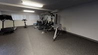 Located at 8795 Via Linda Ave in Whittier, California, this single-story fitness and recreation facility offers a comprehensive layout designed to support both active exercise and casual gathering. The space is divided into distinct functional zones, all unified by durable speckled rubber flooring, neutral gray walls, and textured ceilings illuminated by recessed fluorescent lighting throughout.

The primary workout area is generously proportioned, accommodating a multi-station cable and weight machine at its center, flanked by a stationary bike, elliptical trainer, rowing machine, weight bench, and a treadmill. A large wall-mounted mirror enhances the sense of depth while allowing users to monitor their form. A mounted flat-screen television adds convenience for those working out. Windows with vertical blinds along the perimeter allow natural light to filter in without direct glare.

A secondary section of the fitness area features additional cardio equipment and provides direct access to the outdoors through a set of glass doors, offering ventilation and natural light.

Adjacent to the main gym space is a smaller, carpeted lounge and meeting area furnished with an octagonal wooden dining table and chairs, built-in cabinetry, and a window with vertical blinds, providing a comfortable setting for relaxation or informal gatherings.