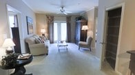 This single-floor space centers around a spacious living area finished with plush, neutral-toned carpeting that adds warmth underfoot. Crown molding lines the ceiling throughout, lending a refined architectural detail that elevates the overall aesthetic. A ceiling fan with light fixture is centrally positioned, providing both air circulation and ambient illumination.

Natural light enters through a set of glass-paned doors dressed with decorative drapery in earthy brown and floral tones, creating a welcoming focal point at the far end of the room. The living area is furnished with a generously sized cream sofa accented with decorative pillows, paired with a classic upholstered accent chair and a glass-top coffee table, creating a comfortable and cohesive seating arrangement.

A large, dark wood armoire anchors one corner of the room, serving as both a storage piece and a statement of traditional style. Additional table lamps provide warm, layered lighting throughout the space. An adjacent entryway or hallway is visible, suggesting a well-connected and functional single-level floor plan throughout.