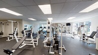 Located at 878 S Oneida St in Denver, CO, the Cedar Run Apartments fitness center is a thoughtfully designed, single-floor facility that provides residents with a comprehensive workout environment. The gym is organized into distinct workout zones, each catering to different training needs.

The strength training area is generously stocked with a full dumbbell rack, multiple weight benches, a Paramount cable machine, a squat/power rack with an attached barbell, and a variety of plate-loaded equipment. Mirrored walls throughout the space enhance the sense of openness and allow for proper form checks during exercise.

The cardio section is equipped with treadmills, including a Spirit brand model, along with a stationary cycling bike, and additional cardio machines positioned near large windows that overlook the surrounding residential community, allowing natural light to fill the space.

Flooring throughout the facility consists of durable rubber gym matting, well-suited for heavy equipment and high-impact workouts. The drop ceiling features recessed panel lighting that ensures consistent, bright illumination across the entire floor. A wall-mounted flat-screen television is also present for entertainment during workouts.

The overall ambiance is clean, functional, and well-maintained, with motivational signage on the walls and clearly marked social distancing indicators on the floor, reflecting a health-conscious and community-oriented environment.
