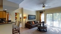 This single-floor residence offers a thoughtfully designed layout that balances comfort and functionality. The living area serves as the heart of the space, featuring plush wall-to-wall carpeting, warm neutral-toned walls, and crown molding that adds a refined architectural touch. A ceiling fan with integrated lighting anchors the room, providing both airflow and ambient illumination. Large windows dressed with decorative curtains allow natural light to filter in, brightening the space and offering a connection to the outdoors. A sliding glass door further enhances the sense of openness.

The living room accommodates generous seating, centered around a round glass-topped coffee table, and is accented by a framed wall art piece that adds a splash of color and personality. A floor lamp provides warm, layered lighting to complement the natural light. An elegant arched doorway connects the living area to an adjacent space, lending a classic architectural character to the interior. The dining area is partially visible, featuring a dining table and upholstered chairs, seamlessly integrated into the overall open flow of the home. The textured ceiling adds subtle dimension, completing the cozy and inviting ambiance of this well-designed residence.