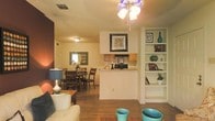 This single-story home features an open-concept layout that seamlessly connects the living room, dining area, and kitchen into one cohesive, functional space. Warm hardwood-style flooring runs throughout, lending a consistent and inviting feel to the interior.

The living area is anchored by a decorative multi-light ceiling fan and offers a comfortable, casual atmosphere. A built-in white bookcase along one wall provides both storage and display space, adding a structured architectural detail to the room. Adjacent to the living space, the dining area accommodates a full dining set beneath a flush-mount ceiling fixture, with a framed mirror on the wall enhancing the sense of openness.

The kitchen features a peninsula-style counter that creates a natural divide between the cooking and living zones while maintaining visual connectivity. Wooden cabinetry, black appliances, and an overhead range hood complete the functional kitchen setup.

Natural light filters into the dining area, brightening the warm, neutral color palette that runs throughout the space. Overall, the layout is practical and efficiently organized, making the most of its single-floor footprint.
