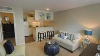 This single-floor unit features an open-concept layout that connects the kitchen and living area, creating a cohesive and functional living space. The living area is finished with plush carpeting in a warm neutral tone, providing a comfortable foundation for the room. A sofa with decorative striped and patterned throw pillows anchors the space alongside a distinctive dark, barrel-shaped ottoman serving as a coffee table.

The kitchen is positioned adjacent to the living space, separated by a raised breakfast bar with seating for two. White cabinetry lines the kitchen walls, complemented by light countertops and standard appliances, including a full-sized refrigerator. The kitchen flooring transitions to hardwood-style material, offering a practical contrast to the carpeted living space. Under-cabinet lighting brightens the work area effectively.

The walls throughout are finished in a soft, warm gray tone, providing a neutral backdrop. Two framed green and blue prints hang above the kitchen pass-through, adding a subtle decorative accent. The textured ceiling features a flush-mount light fixture. Overall, the space offers a clean, well-organized layout with a cohesive neutral palette suited for comfortable everyday living.